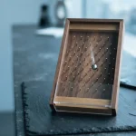 A wooden Plinko board with a steel ball beside a blurred phone and payment cards on a slate counter