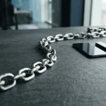 Polished steel chain curving across a slate table, with a smartphone, hardware wallet and payment card in a row