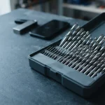Polished steel drill bits in an open fitted case, with a hardware wallet and smartphone behind