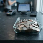 Unmarked silver and copper coins in a glass tray on a slate desk, with a hardware wallet beside a modern cashier counter