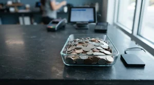 Unmarked silver and copper coins in a glass tray on a slate desk, with a hardware wallet beside a modern cashier counter