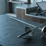 Two crossed fencing foils on a slate tabletop beside a softly blurred phone, hardware wallet and metallic coins