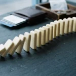 Line of plain dominoes toppling across a slate table, with payment cards sliding from a holder beside a cashier tray