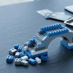 Half-built bridge of plain interlocking toy bricks on a slate table, with payment cards sliding toward a cashier tray