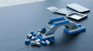Half-built bridge of plain interlocking toy bricks on a slate table, with payment cards sliding toward a cashier tray