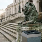 Bronze lion statue on a stone pedestal above civic steps, with a smartphone and hardware wallet on a kiosk counter