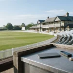 Sunlit cricket pavilion and empty pitch behind a brushed-metal payments counter with a phone, wallet and loose coins.