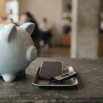 Pale blue ceramic piggy bank on a slate counter, with a smartphone and hardware wallet on a cashier tray
