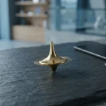 A polished golden spinning top on a slate tabletop, with a phone and loose payment cards in soft daylight behind it