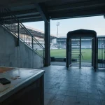 Daylit stadium turnstile overlooking an empty football pitch, with a small hardware wallet and plain coins on a nearby counter