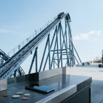 A steel roller coaster climbing a steep track, with a phone and unmarked coins at a nearby cashier counter