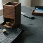 Wooden dice tower with two brass dice tumbling into a felt tray, with a hardware wallet and payment cards behind